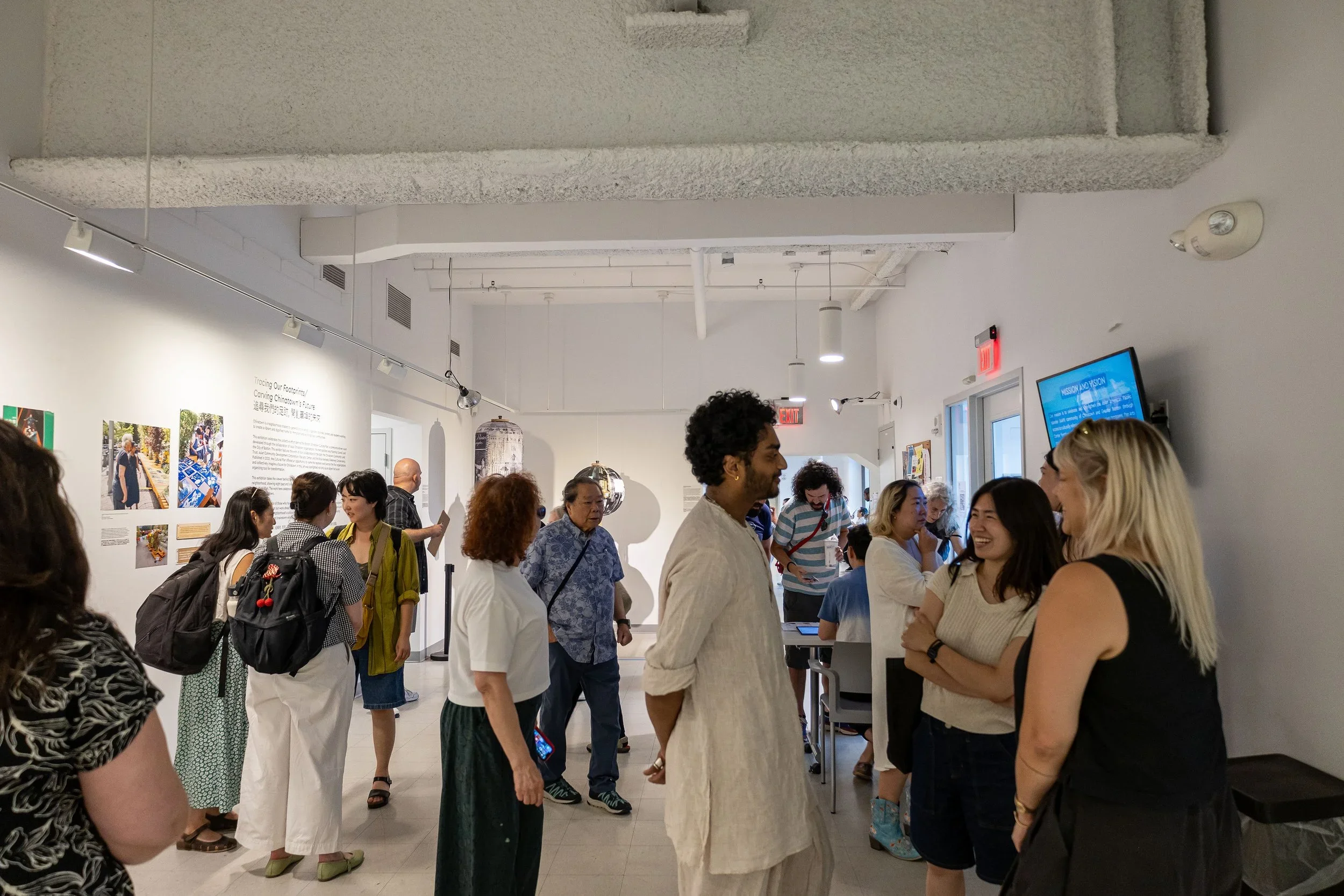 A group of people gathered inside an art gallery or museum, engaged in conversations, with some looking at displays or listening to a speaker. The room has white walls, hanging lights, and informational posters about an exhibit.