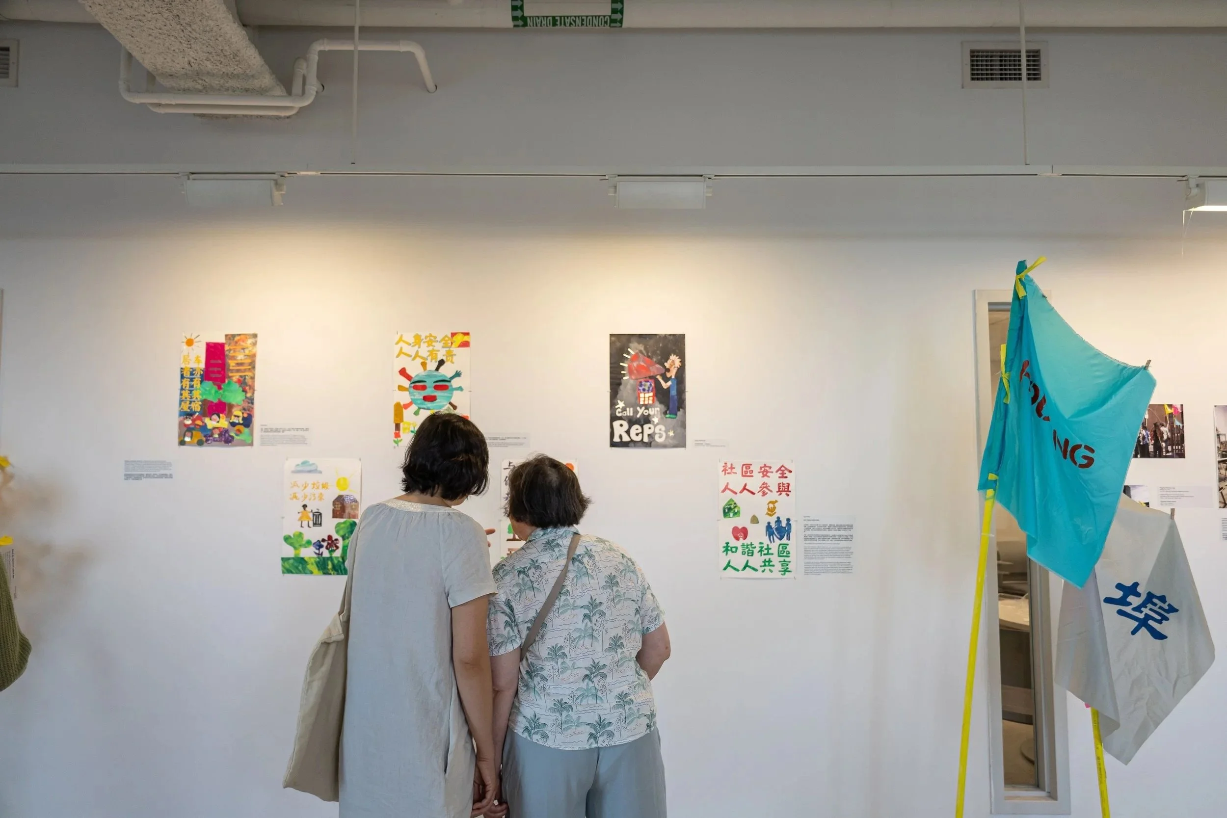 Two women stand in front of a white wall with colorful posters and signs, some featuring Chinese characters. The women are engaged in conversation or looking at the posters, and there is a blue flag on a yellow pole to the right.