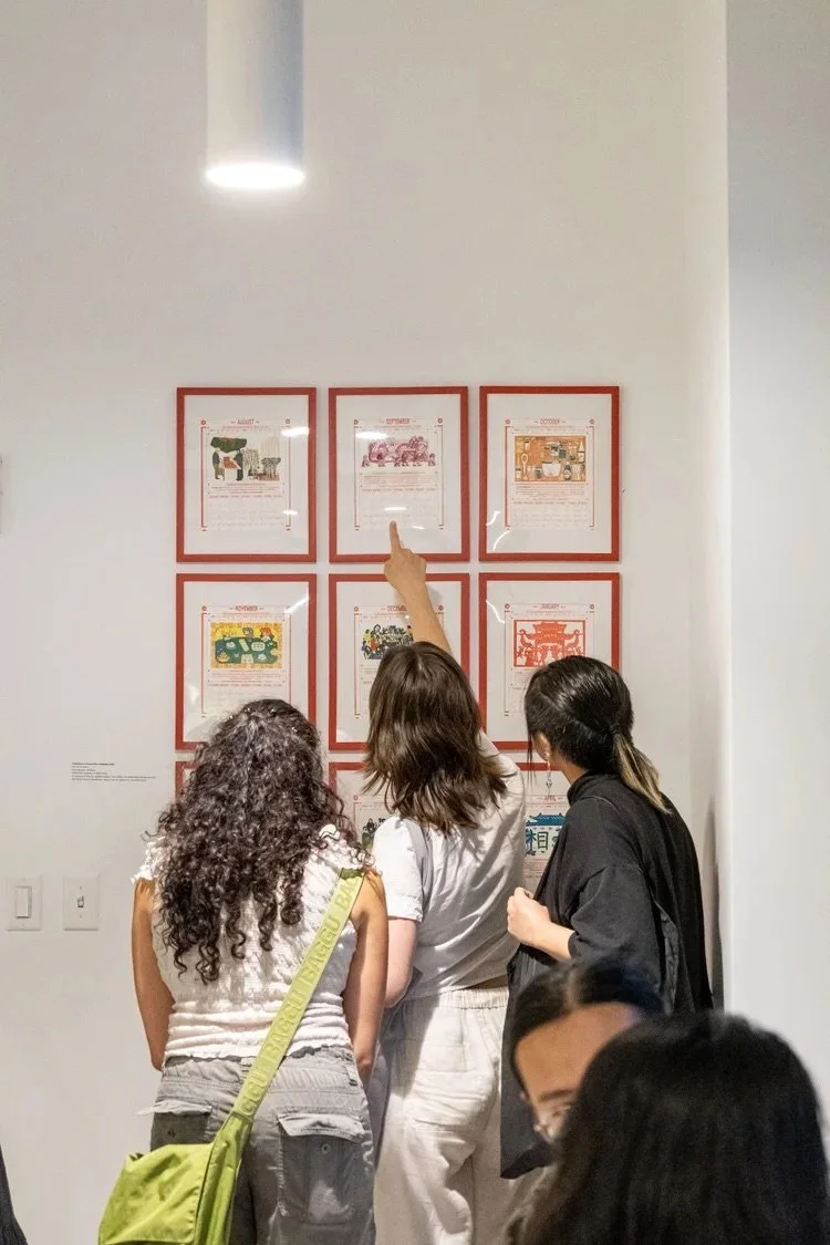 Three people looking at framed pictures on a white wall in an art gallery, with one pointing at the middle picture.