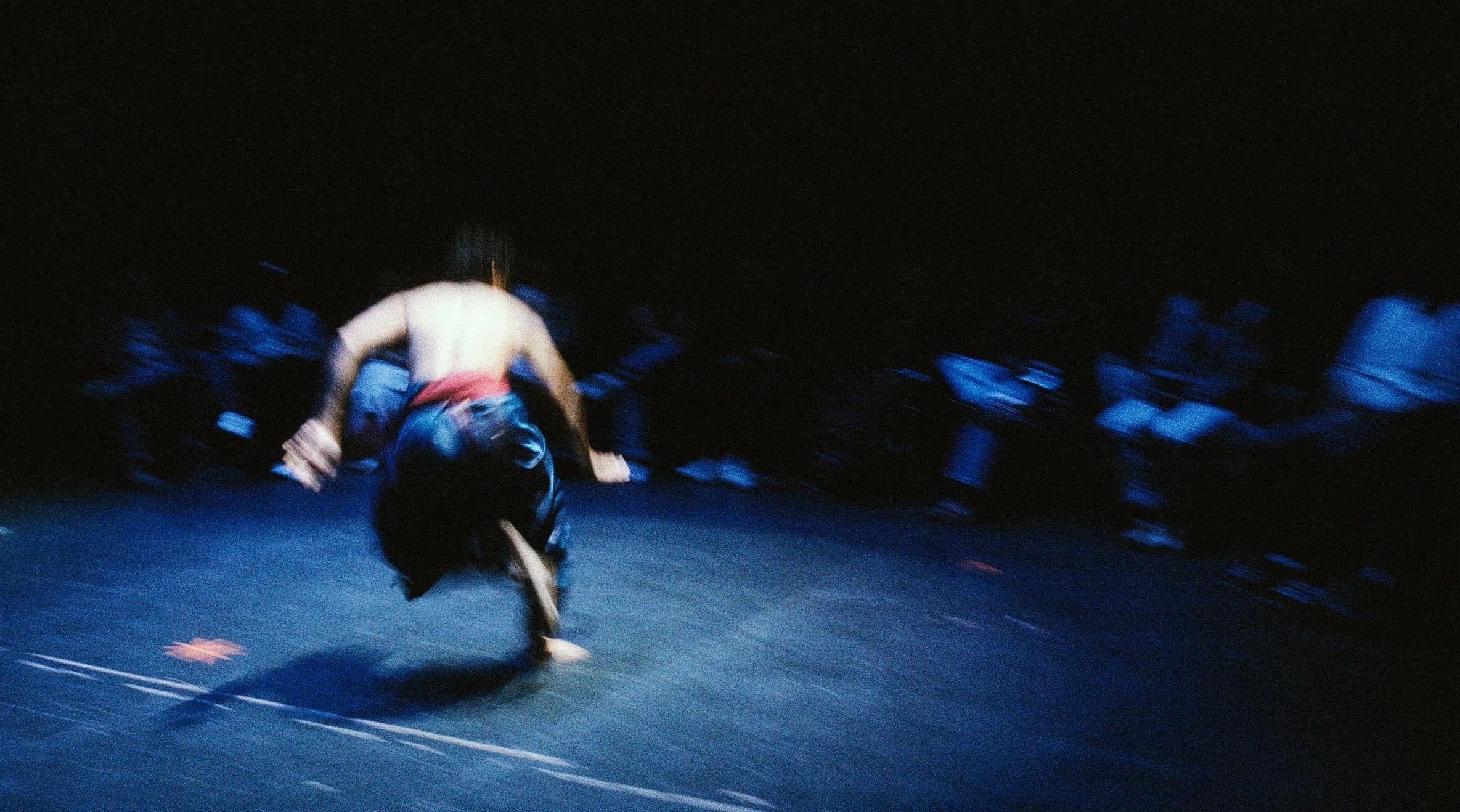 A person performing a handstand or hand walk on a dark stage, with the background filled with an audience or fellow performers watching. The scene appears to be in motion, creating a dynamic and energetic atmosphere.