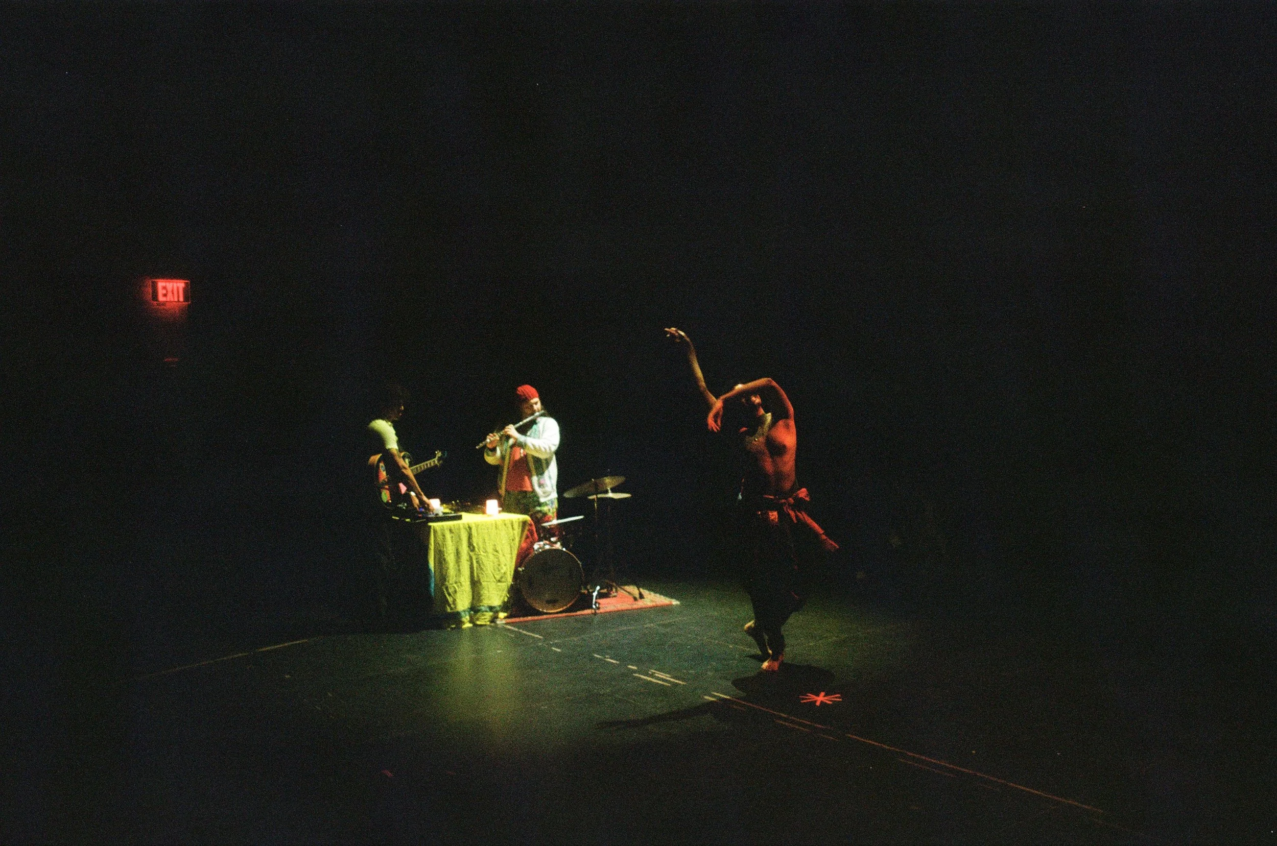 A dark stage with three performers. One person is playing a guitar, another is playing a flute, and the third is dancing. There is a yellow table with equipment, a drum, and a red star-shaped object on the floor. An illuminated exit sign is visible i