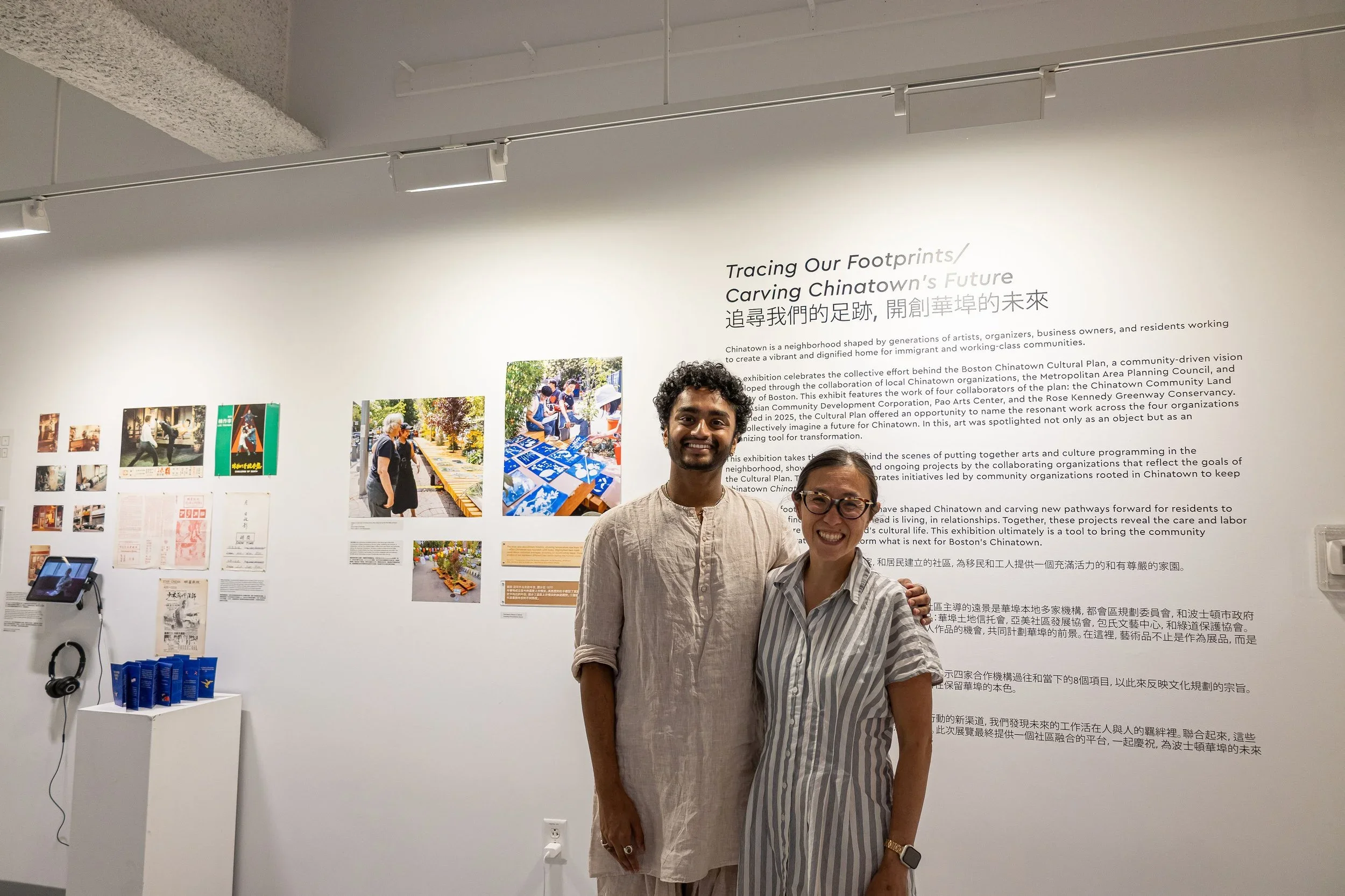 Two smiling individuals, one man and one woman, standing together at an indoor exhibition with a white wall behind them. The wall features text titled "Tracing Our Footprints / Carving Chinatown's Future" and displays photographs and informational po