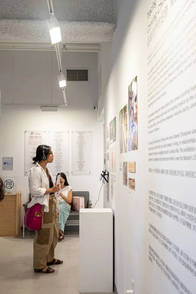 Woman with headphones, standing and reading a brochure at an art exhibit, with framed pictures and text on the wall, and a woman sitting on a bench in the background.