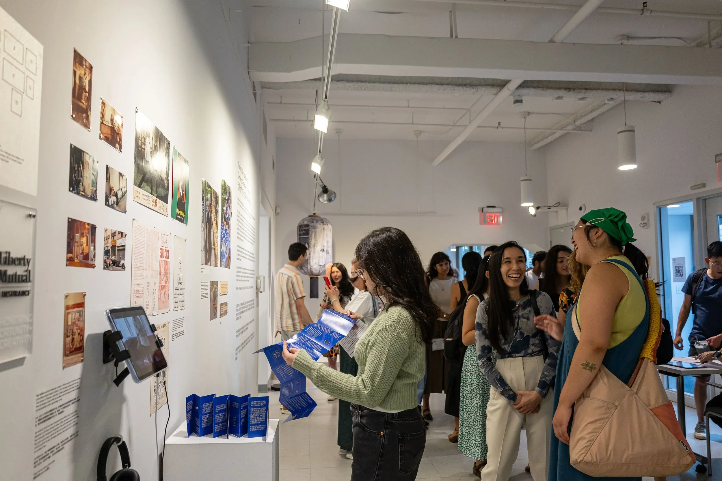 A group of people inside a gallery or museum, looking at displayed artworks and informational panels on the wall. Some are engaging with brochures or tablets, and the ceiling has exposed pipes with track lighting.