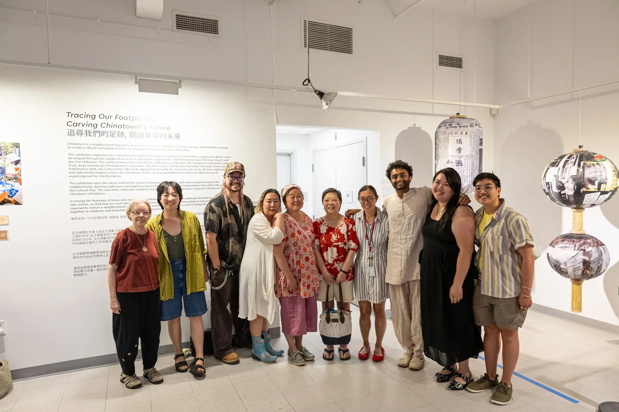 Group of diverse people standing together in an art gallery, smiling for a photo, with informational text on the wall behind them.