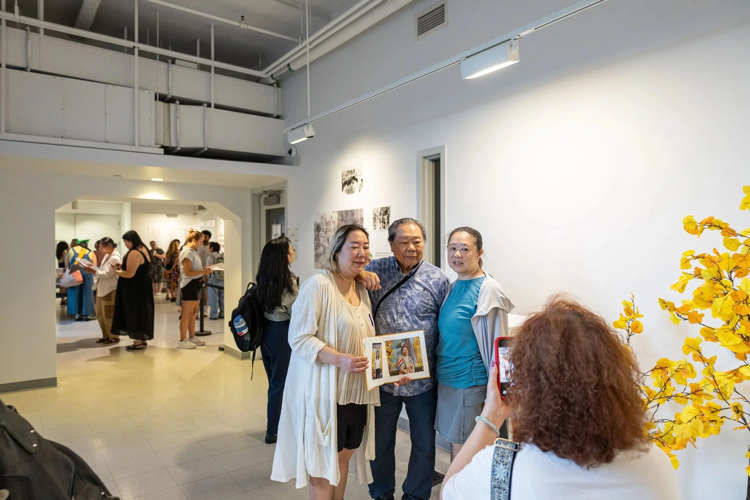 A group of four people standing together at an art exhibit, with various art pieces hanging on the white walls in the background. Other visitors are seen browsing and engaging with the artwork in the space.