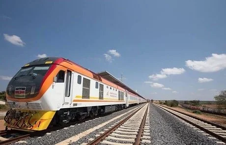 A modern train on railway tracks in a rural area under a clear blue sky.