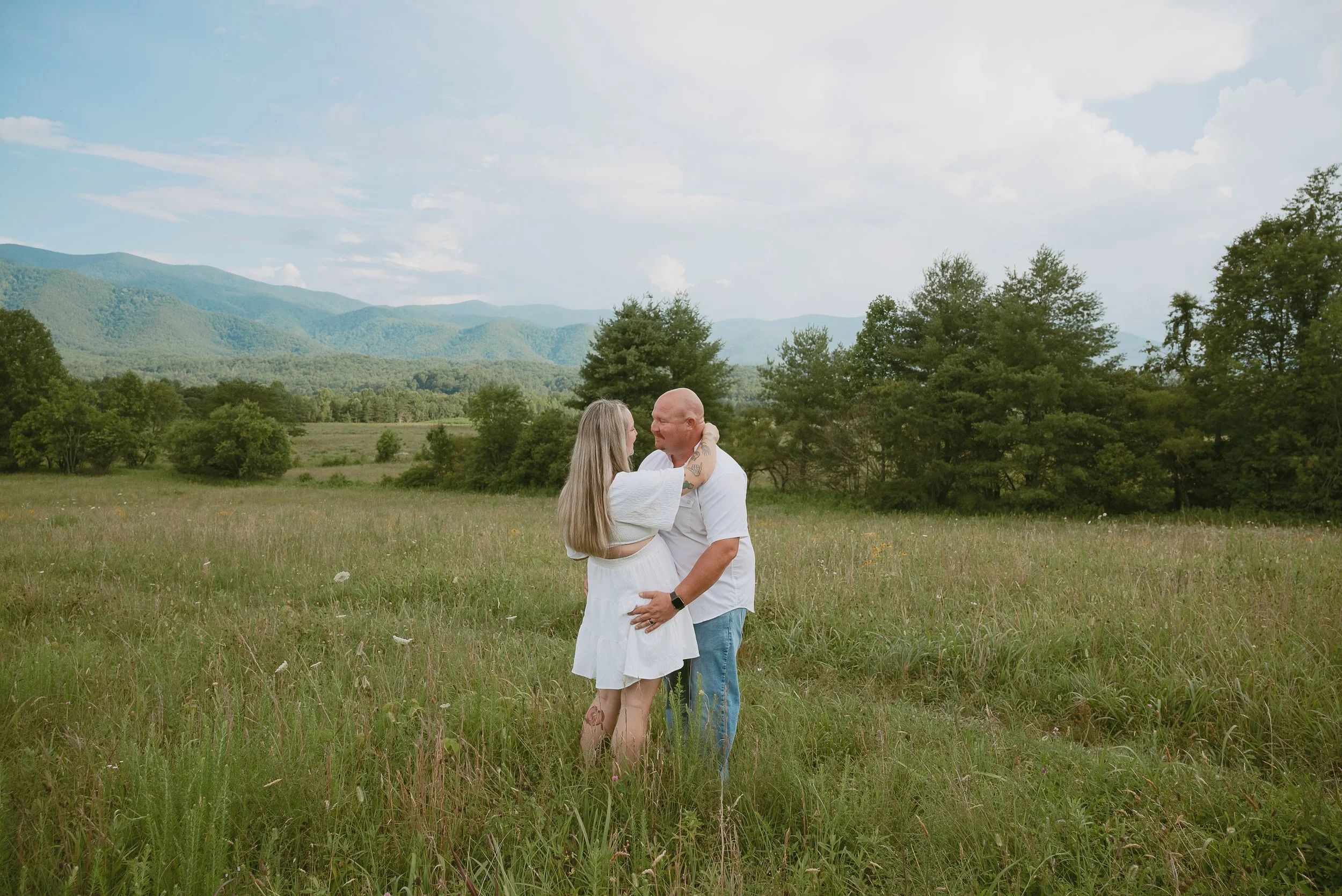 A couple standing close together in a grassy field, looking at each other with smiles. The woman has long blonde hair and tattoos on her leg, while the man is bald and has tattoos on his arm. There are trees and mountains in the background, under a partly cloudy sky.