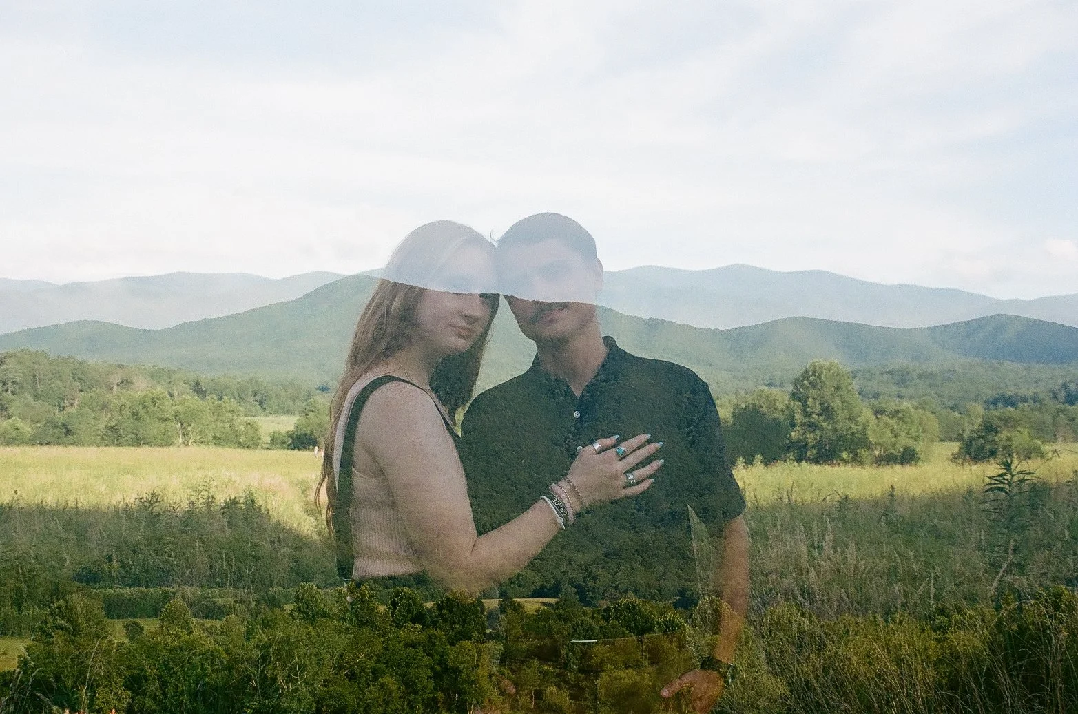 A double exposure photograph of a young couple standing in front of a scenic landscape with rolling hills and trees, the woman's hand resting on the man's chest.