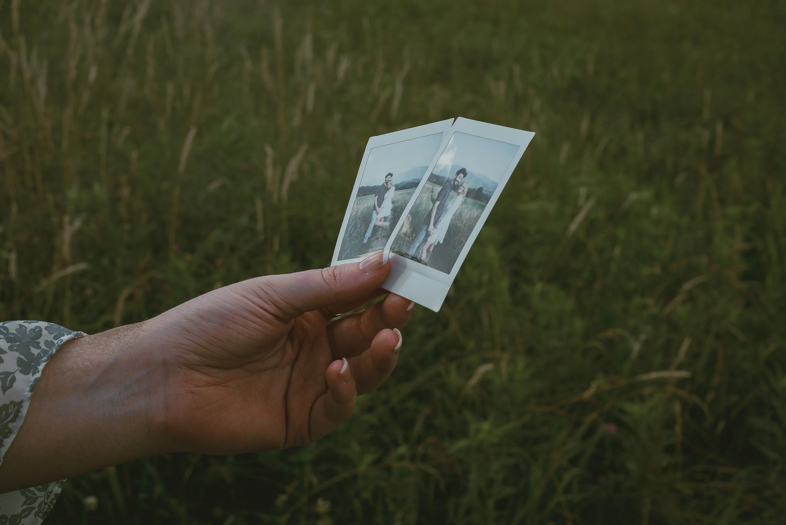 A person holding two instant photographs in front of a grassy field, with a woman in a white dress in the photos.