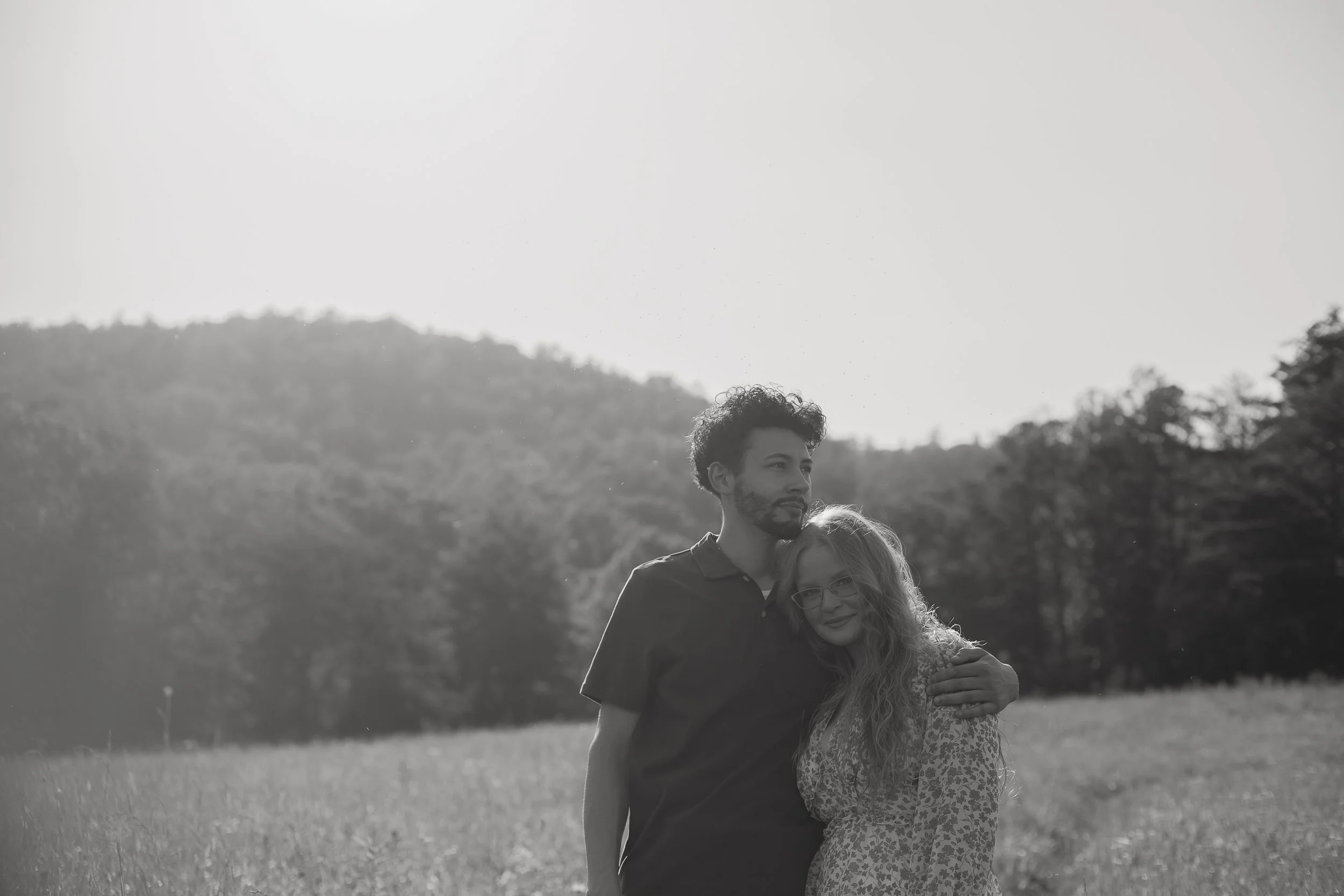 A young man and woman standing together outdoors in a grassy field with trees and mountains in the background, embracing each other.