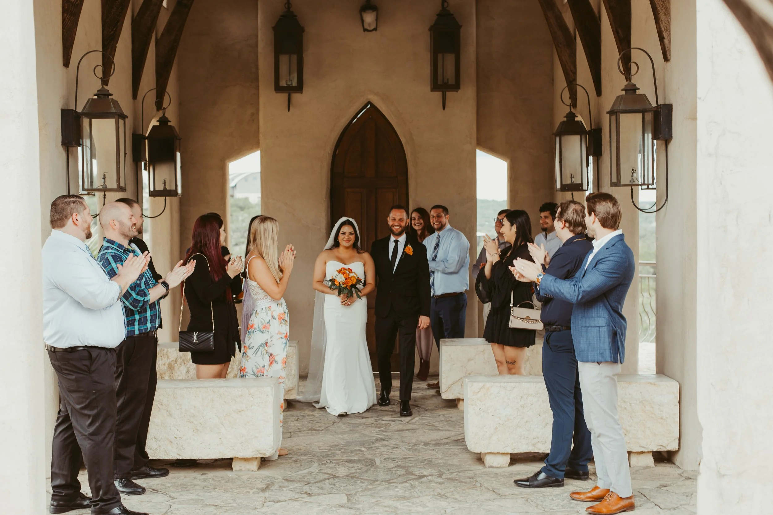 Bride and groom walking down the aisle together, surrounded by clapping friends and family, in an indoor wedding ceremony with rustic decor.