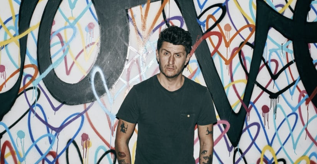 A young man with dark hair and tattoos on his arms standing in front of a colorful graffiti wall.