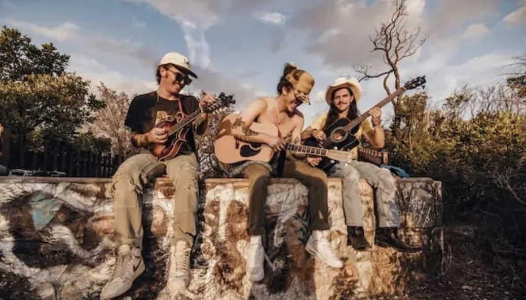 Three people sitting on a brick wall playing guitars outdoors, with trees and a partly cloudy sky in the background.