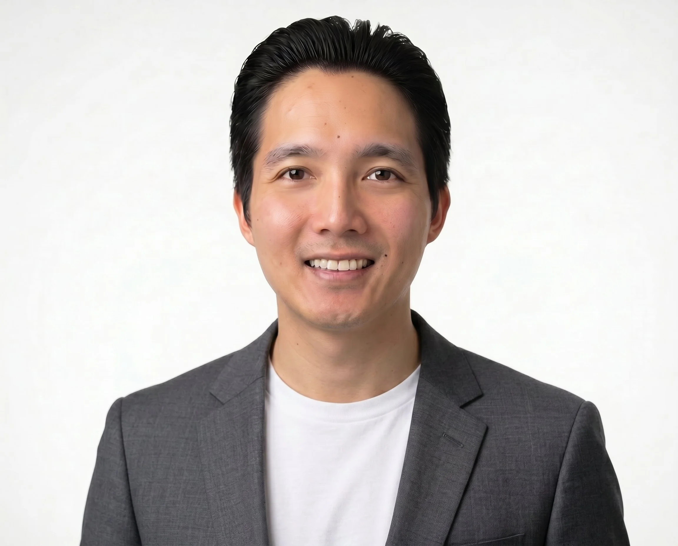 A young man with short black hair, smiling, wearing a dark gray suit jacket and white t-shirt, standing against a plain white background.