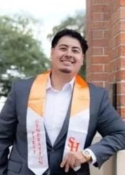 Young man in a suit with graduation stole smiling outdoors near a brick wall
