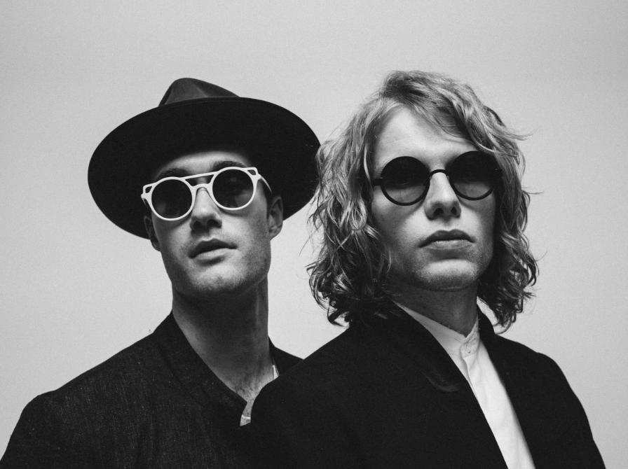 Two people wearing sunglasses and stylish clothing, posing against a plain background.