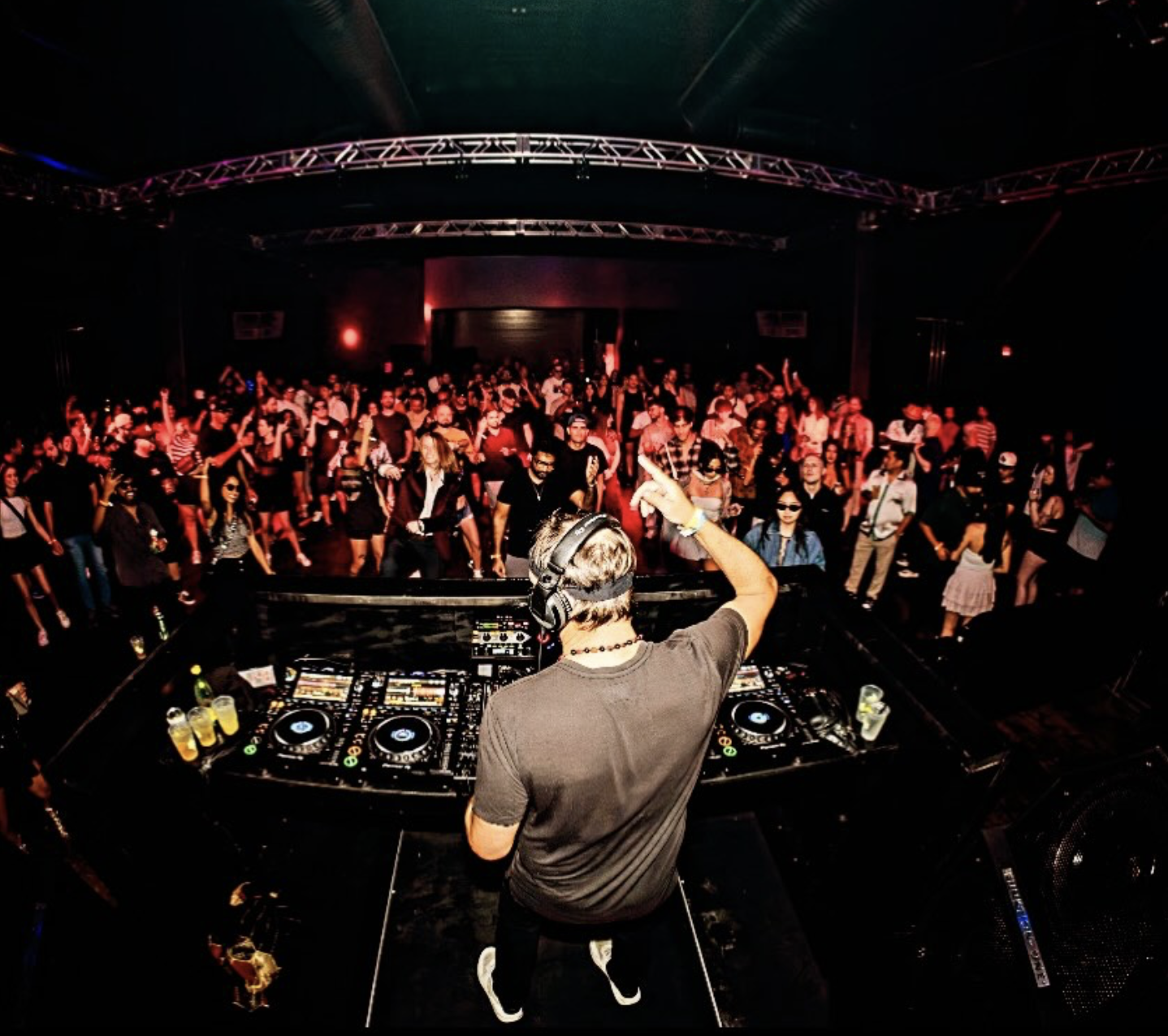DJ performing at a club with a large crowd dancing on the floor, colorful lighting, and music equipment in the foreground.