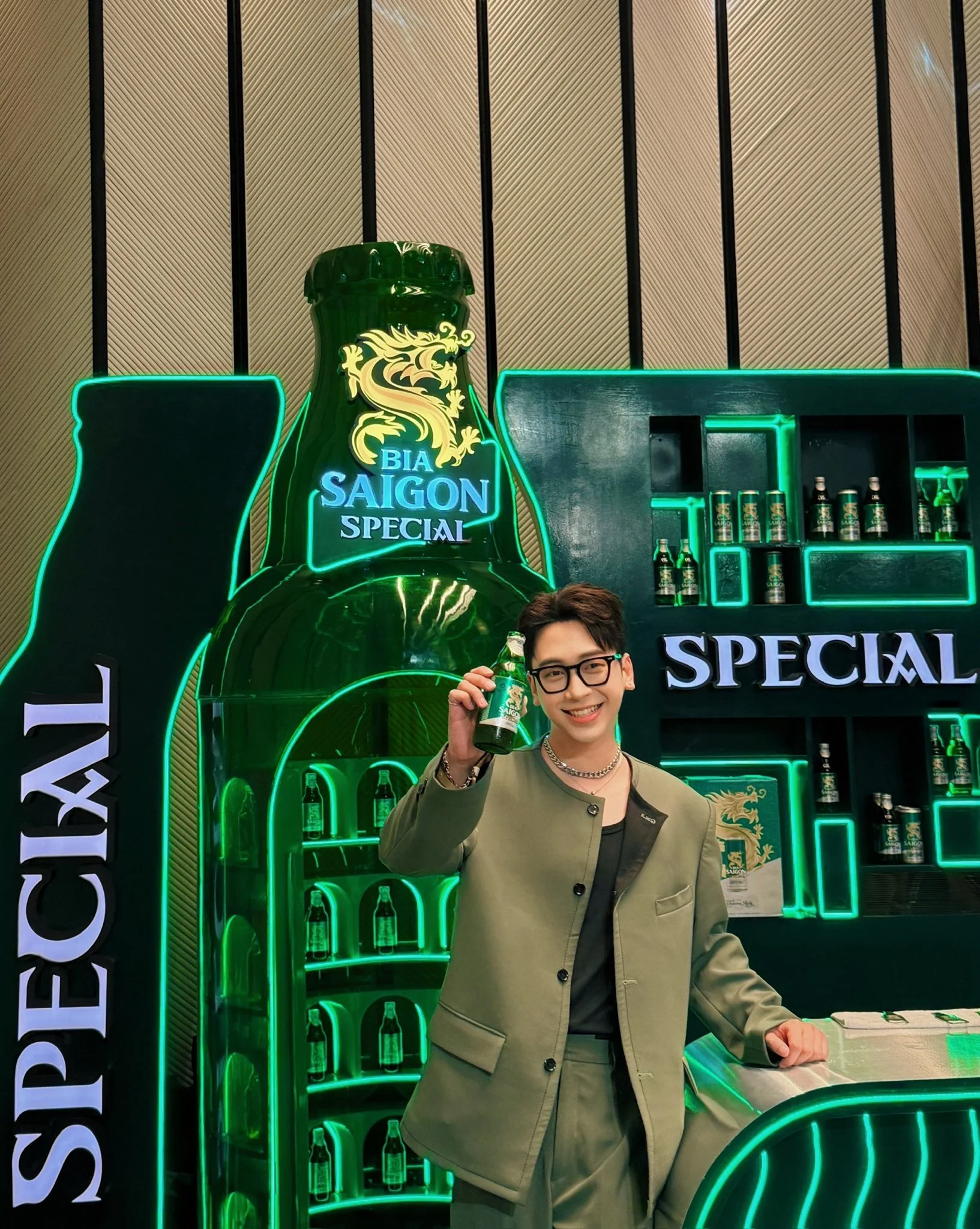 A young man with glasses and a chain necklace holding a bottle of Bia Saigon Special beer in front of a neon-lit bar setup with bottles displayed in the background, and neon signs reading 'SPECIAL' and 'SAIGON'.