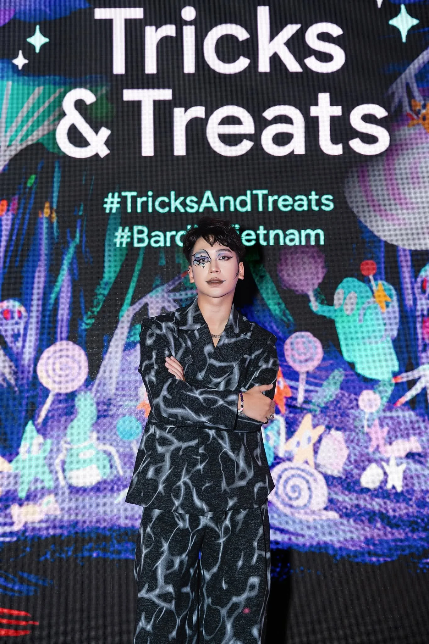 A person with short black hair and artistic face makeup standing with arms crossed in front of a colorful digital screen displaying promotional text for Tricks & Treats event.