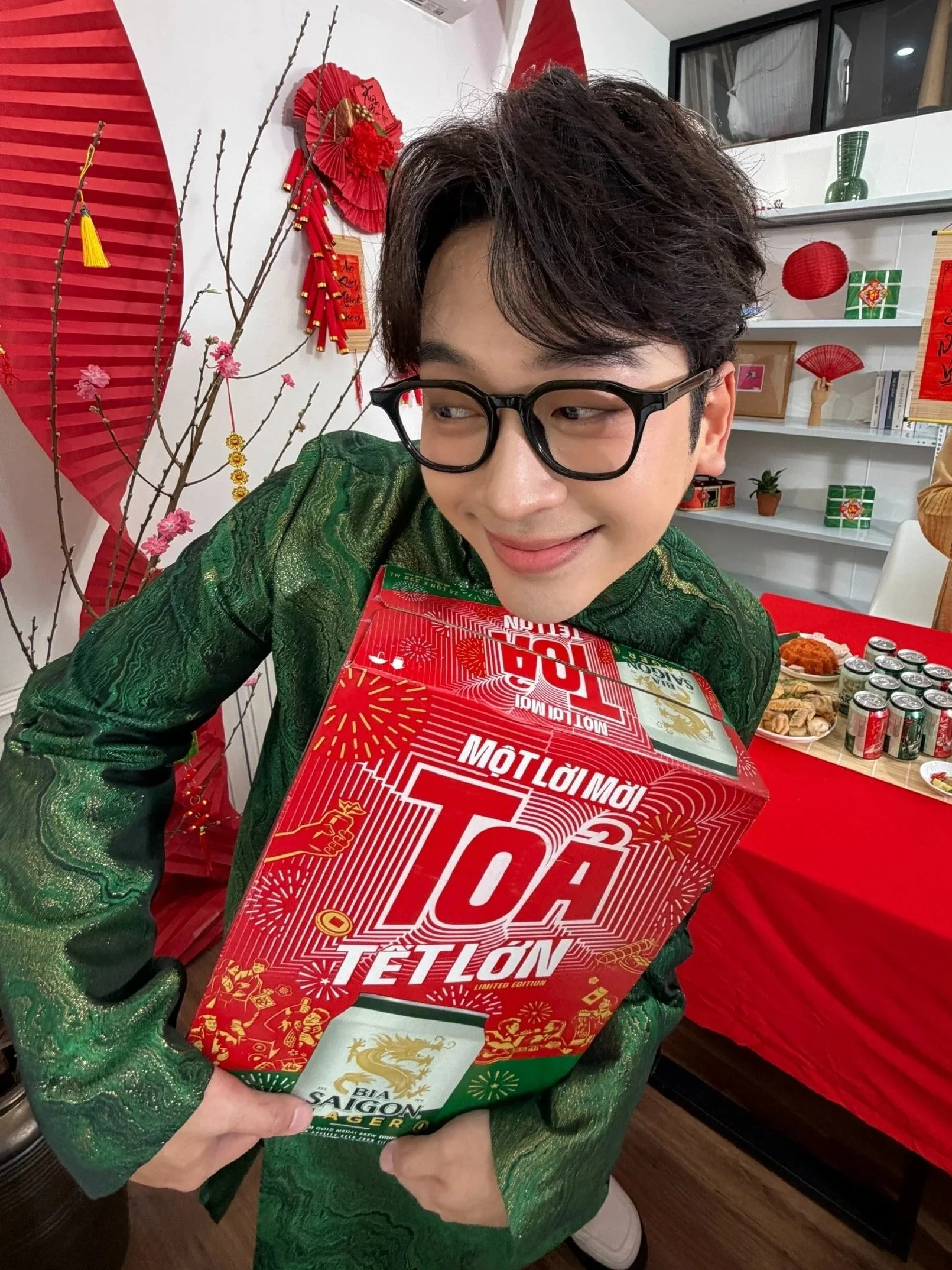 A man with glasses and dark hair wearing a green patterned shirt holding a large red box of Tet Nguyen Limited Edition Tet Lot. The background has red and white decorations including branches with pink flowers, fans, lanterns, and a shelf with books, a small plant, and decorative items.