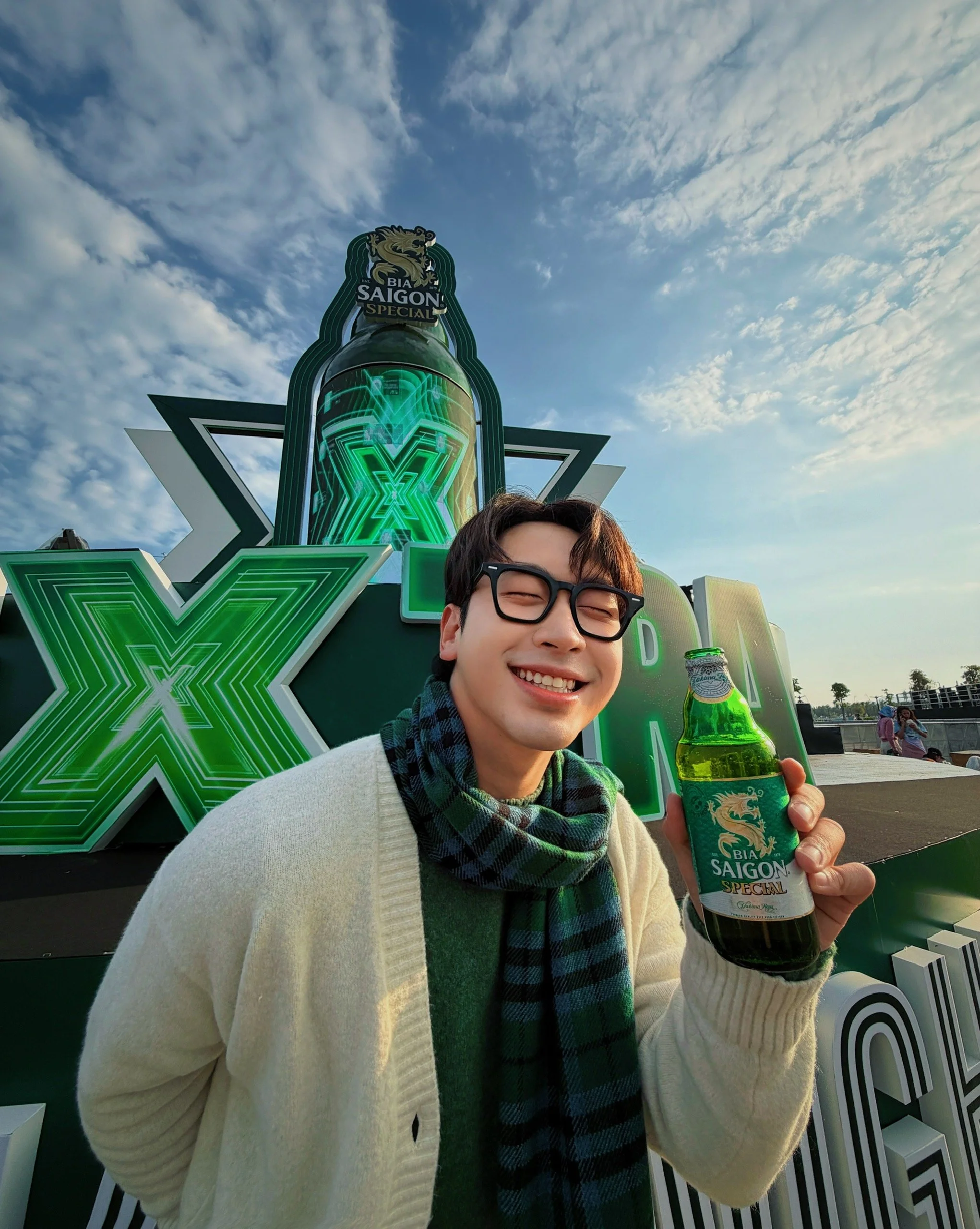 A young man smiling and holding a green bottle of Saigon Special beer. He is wearing glasses, a beige cardigan, and a black and green checkered scarf. In the background, there is a large neon sign with the letter X and a logo of a lion, with the sky and some trees visible.