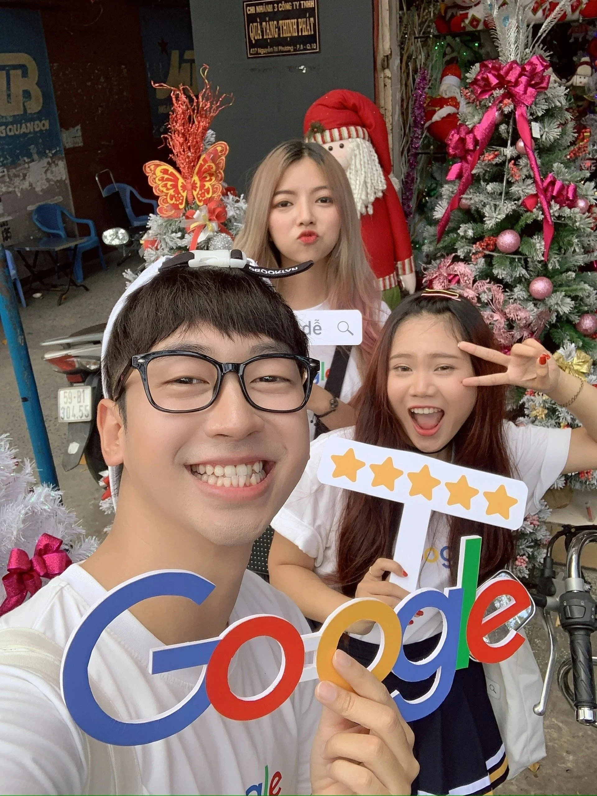 A group of three young people taking a selfie at a festive holiday setting, with Christmas decorations including a decorated Christmas tree and Santa Claus figure in the background. They are holding colorful signs featuring the Google logo and stars.