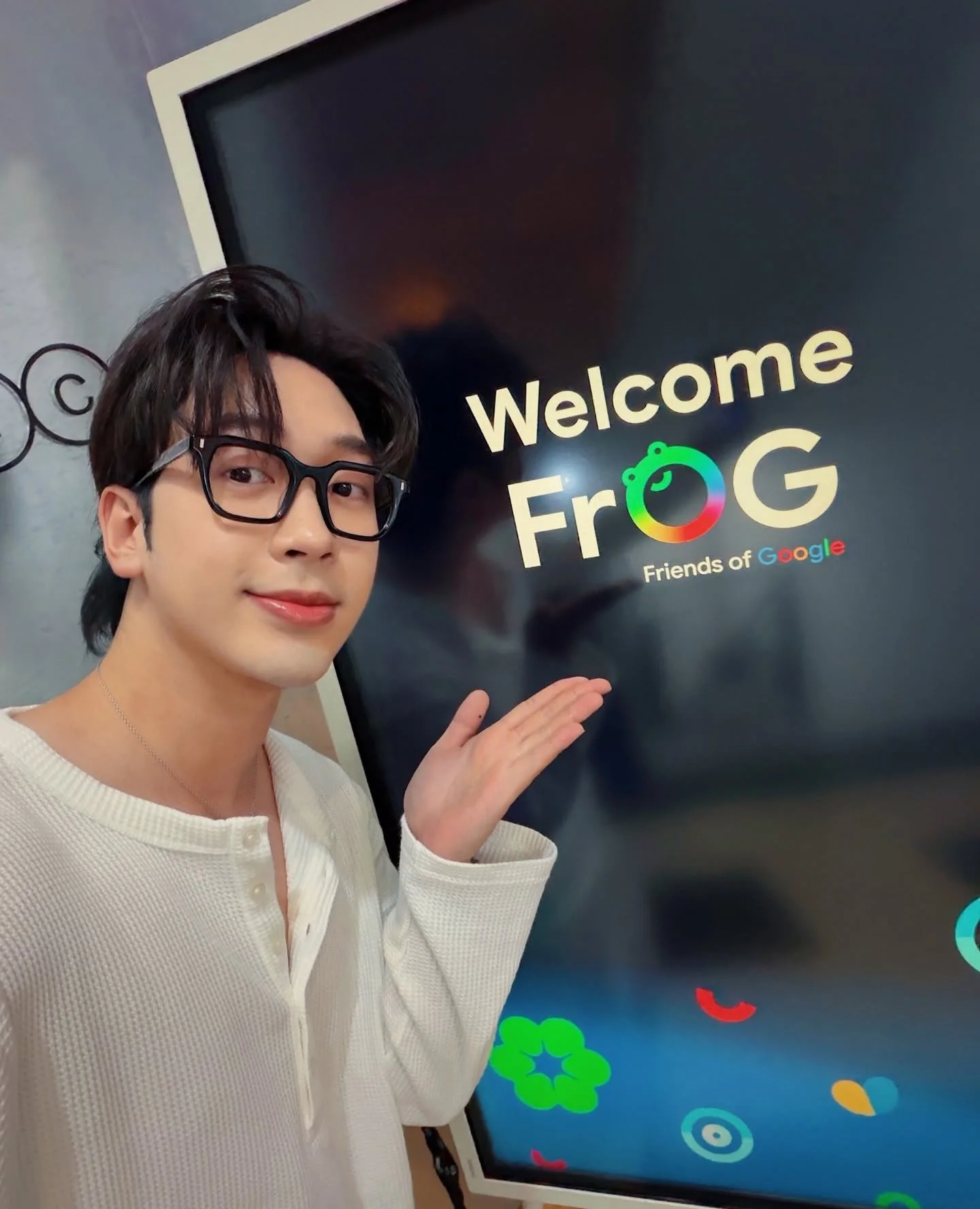 A young man with dark hair, glasses, and a white cardigan, standing indoors in front of a large screen that reads "Welcome FroG Friends of Google." The screen has colorful graphics.