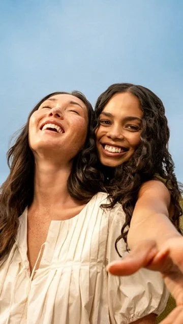 Two women taking a selfie outdoors against a blue sky, smiling and laughing.