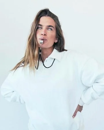 A woman with long, wavy hair wearing a white sweatshirt has a whistle in her mouth, with one hand on her hip and the other hanging by her side, against a plain white background. Hair and makeup by Amber Rose.