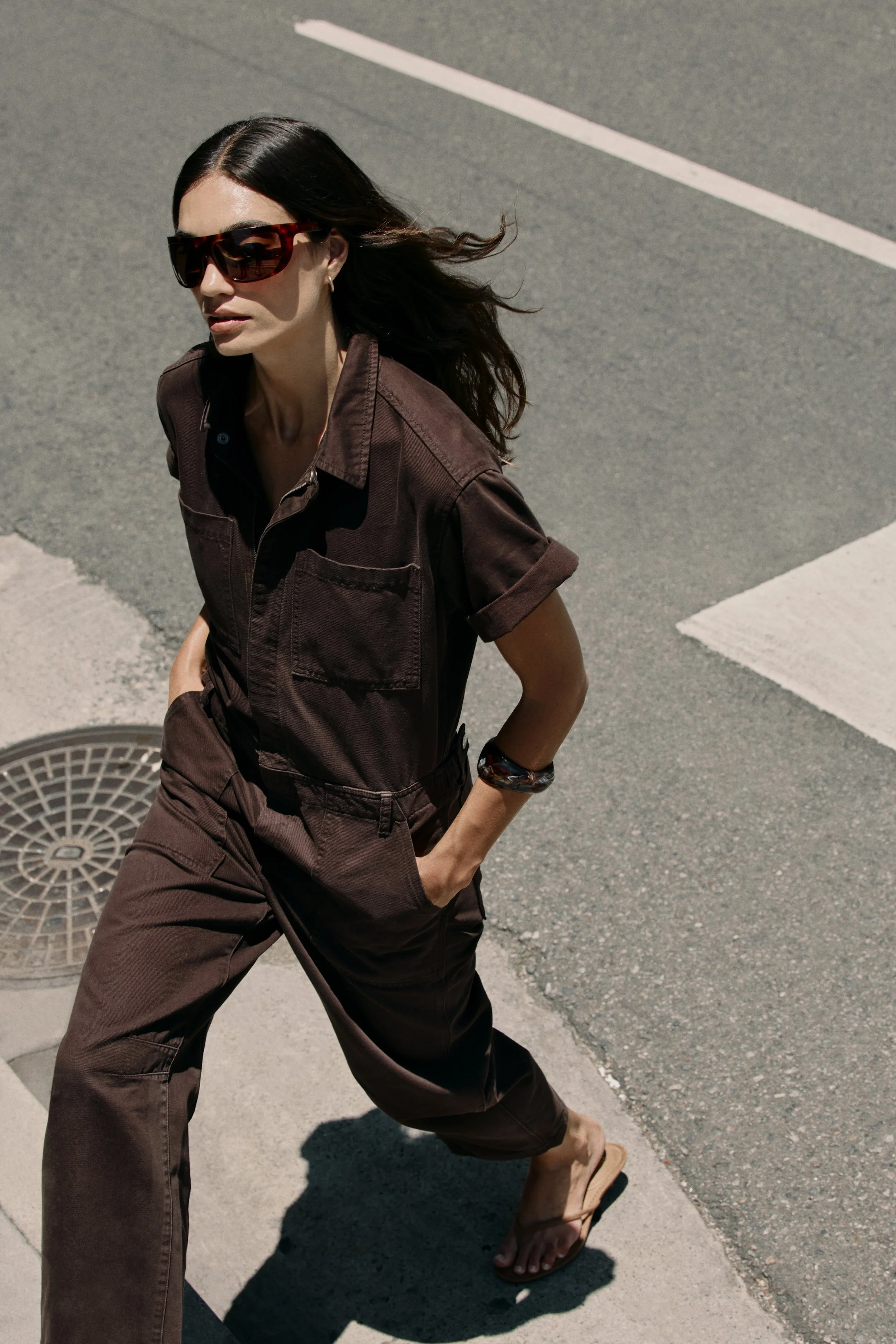 A woman in a brown jumpsuit walking barefoot on an urban street with her hands in her pockets, wearing large sunglasses and a bracelet, casting a shadow on the pavement.