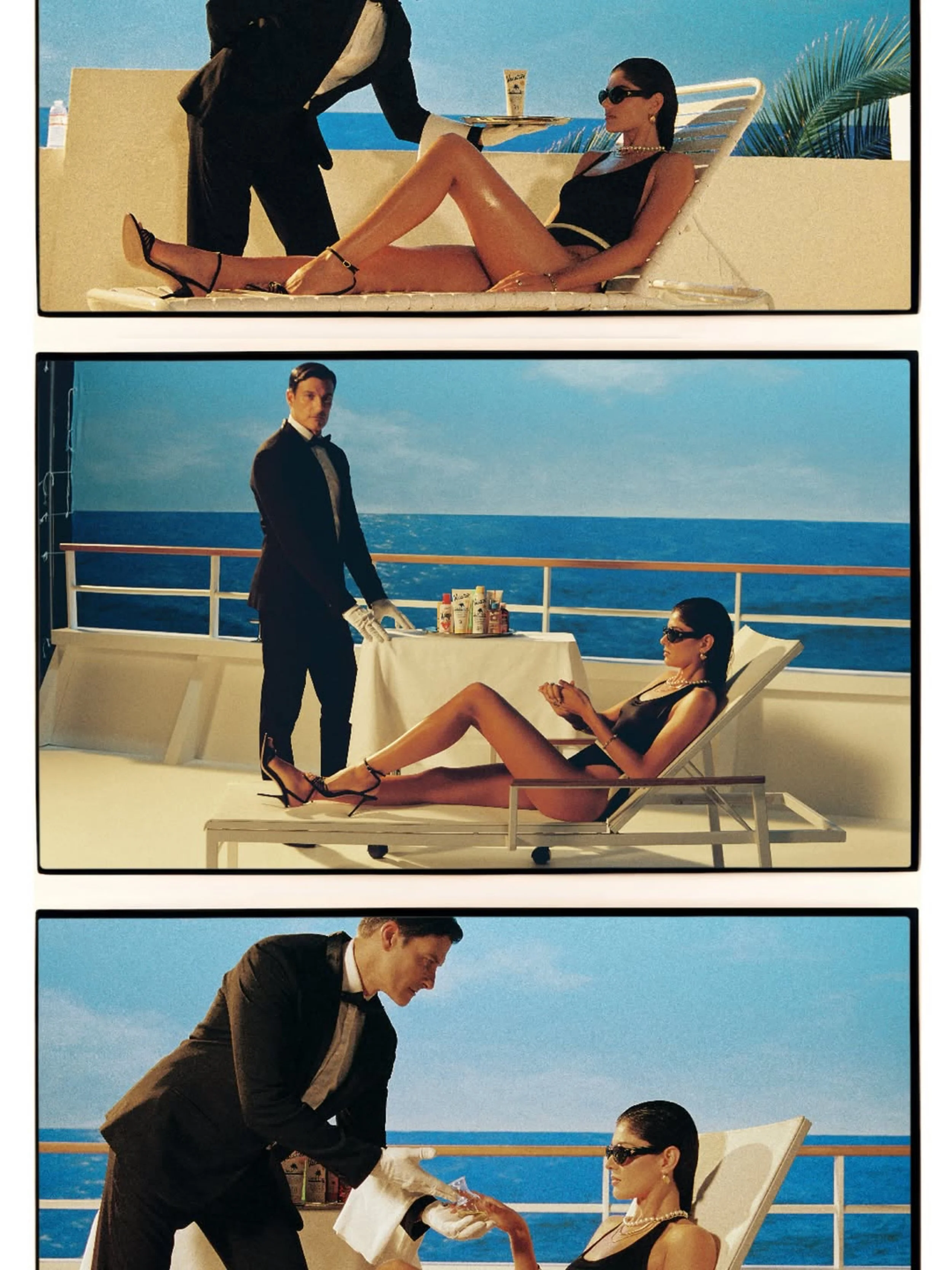 A woman in black swimsuit and sunglasses reclining on a lounge chair on a cruise ship deck, being served drinks by a waiter in a tuxedo. The ocean is in the background. Hair and makeup by Amber Rose.