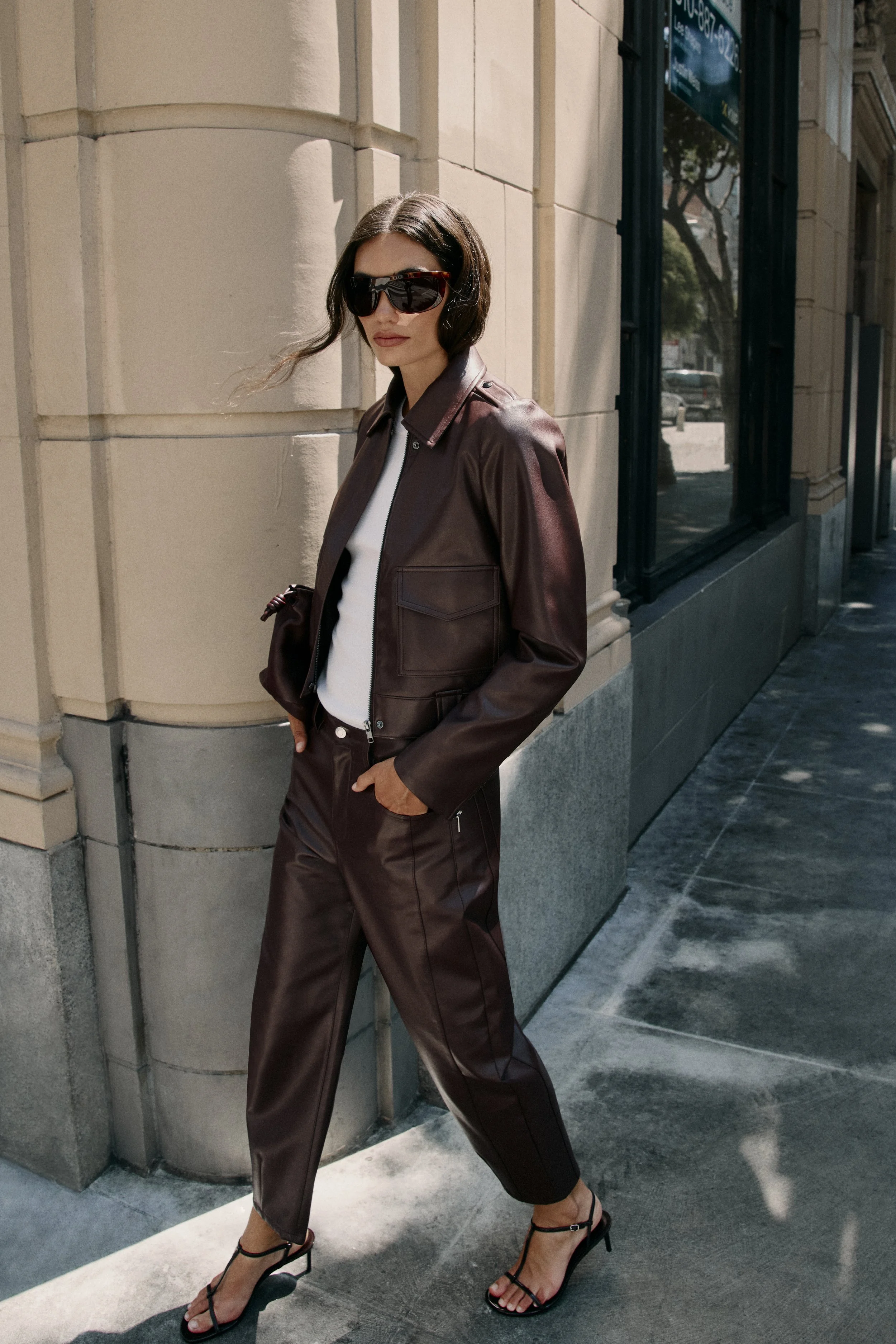 A woman with dark hair wearing oversized sunglasses, a brown leather jacket, and matching leather pants, standing on a sidewalk against a beige stone building with large windows. She is wearing high-heeled sandals and has one hand in her pocket, look