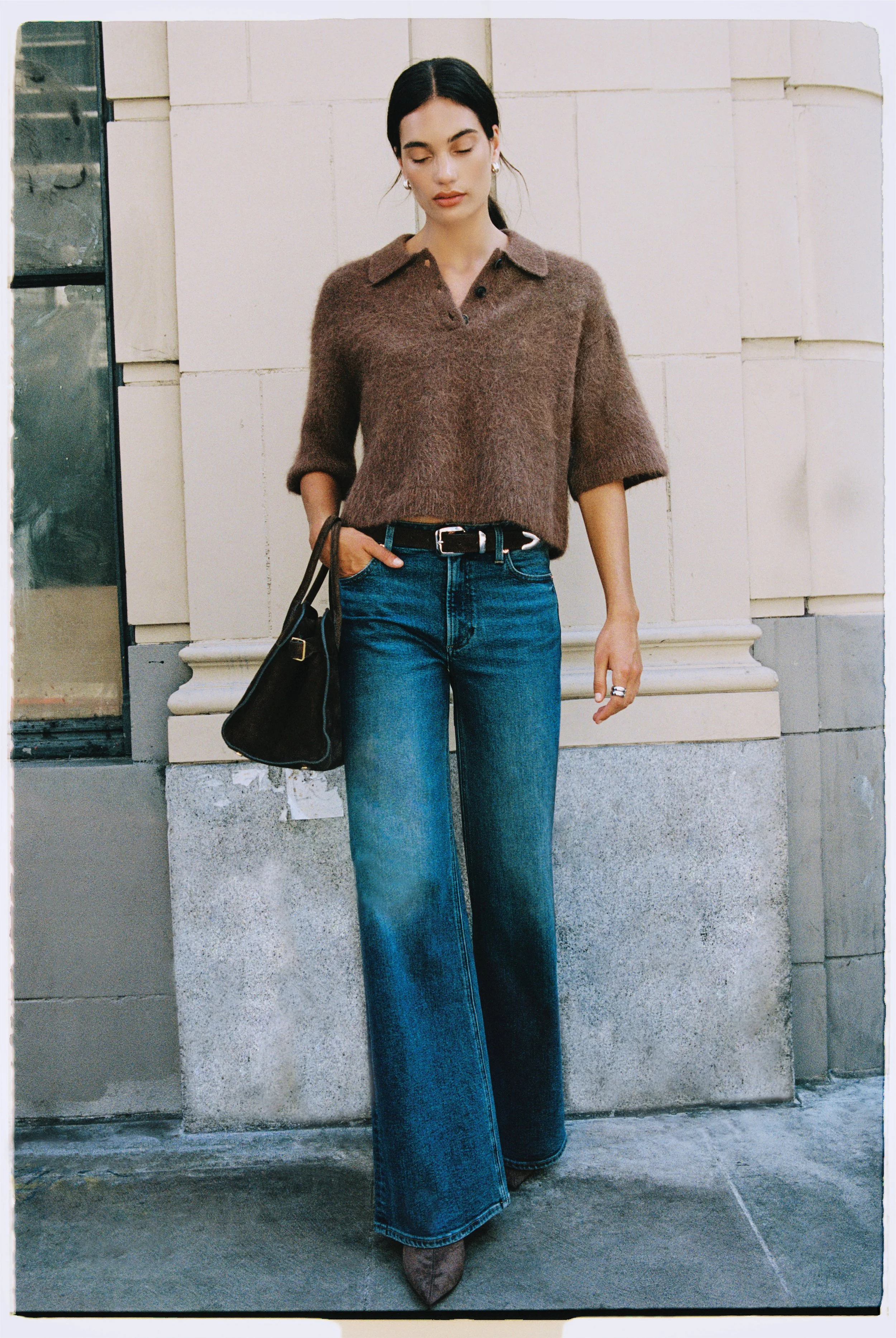 A young woman with dark hair, closed eyes, and light skin, wearing a brown fuzzy sweater with rolled-up sleeves, high-waisted wide-leg jeans, brown pointed shoes, and carrying a black handbag, standing against a light-colored stone wall exterior.