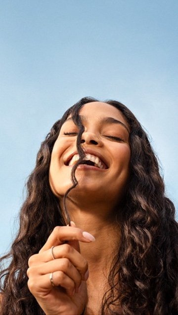 A woman with curly hair smiling and holding a lock of her hair in front of her face.