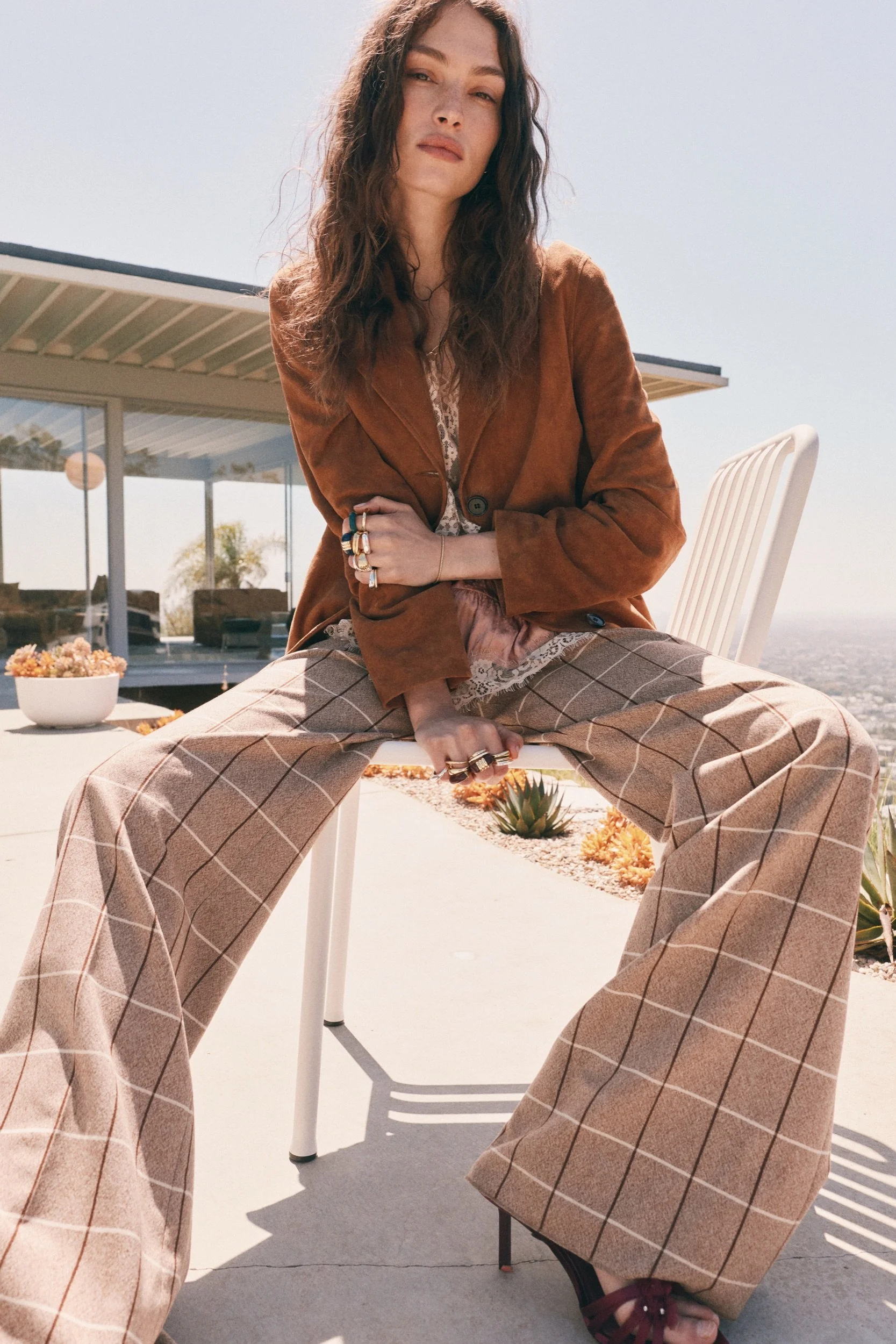 A woman in a brown blazer and plaid wide-leg pants sitting outdoors on a white chair, with a modern house, plants, and a clear sky in the background.