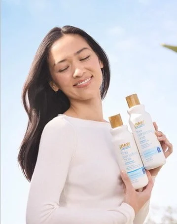 A woman with long dark hair smiling outdoors while holding two bottles of skincare or sunscreen products.