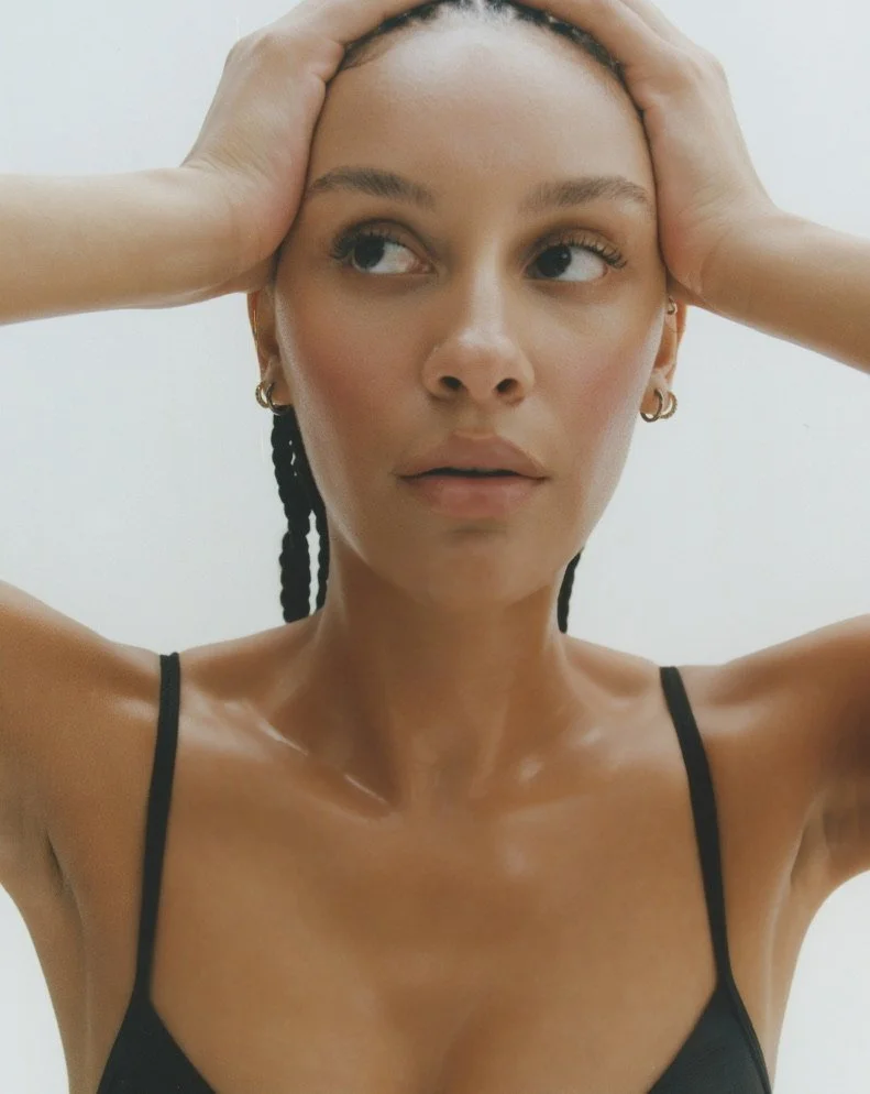 A woman with dark braided hair, wearing a black spaghetti strap top, holding her head with both hands, looking to the side. Hair and makeup by Amber Rose.