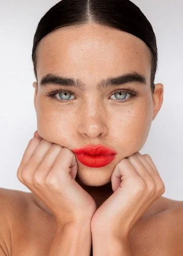 Close-up of woman with dark hair, blue eyes, and red lipstick, holding her face with her hands. Hair and makeup by Amber Rose.
