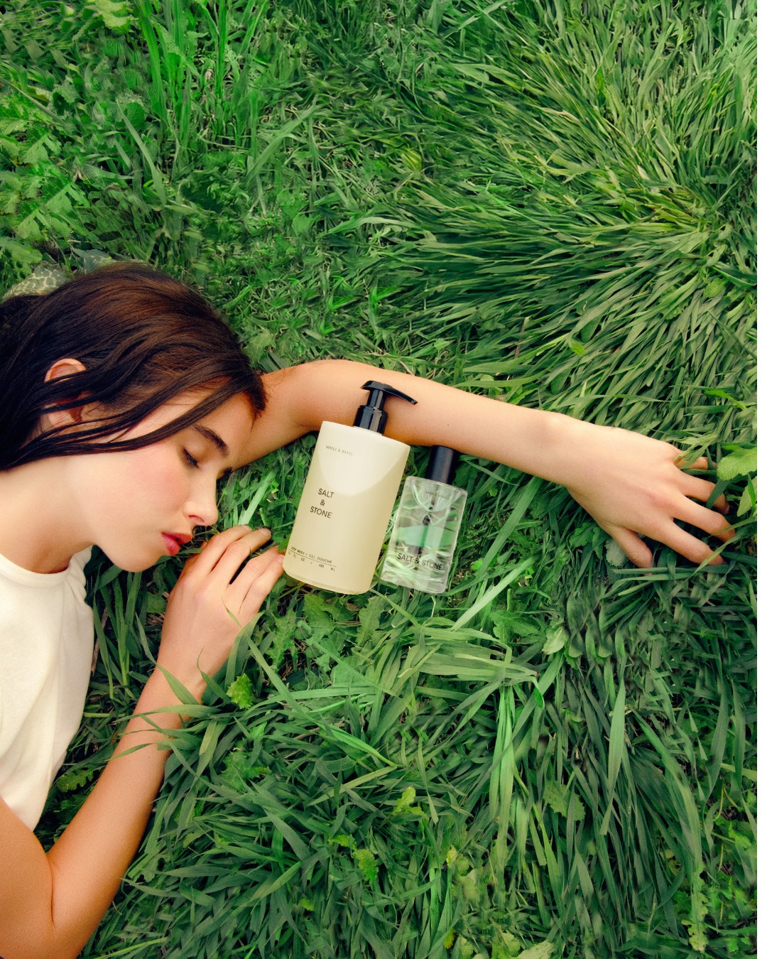 Young woman with dark hair lying on green grass, surrounded by plants, with two bottles of Salt & Stone skincare products nearby.
