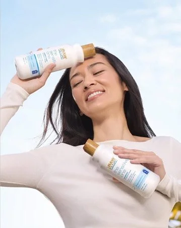A woman smiling outdoors holding two bottles of sunscreen, one near her forehead and one near her chest.