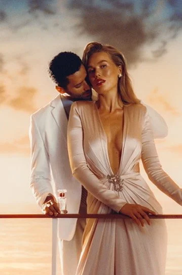 A man in a white suit and a woman in a glamorous beige dress standing close together on a balcony with a sunset sky in the background. Hair and makeup by Amber Rose.