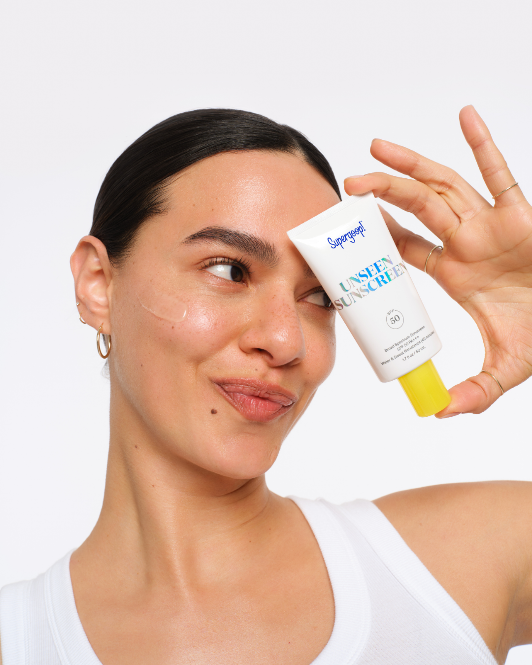 A woman with dark hair and earrings holding a tube of sunscreen near her temple. Hair and makeup by Amber Rose.