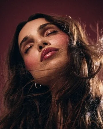 Close-up of a woman with long wavy brown hair, eyes closed, smiling softly, against a dark background. Hair and makeup by Amber Rose.