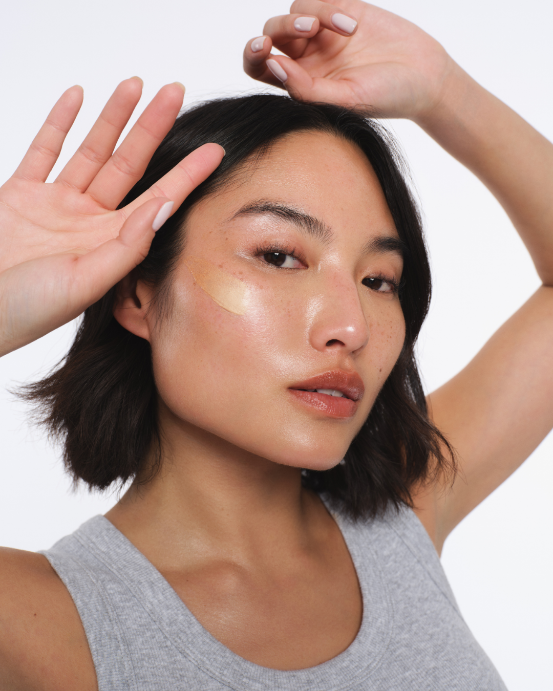 A woman with short dark hair and fair skin showing her face, with one hand raised near her hair and the other near her face. She has clear, glowing skin and is wearing a gray sleeveless top. Los Angeles premiere hair and makeup artist, Amber Rose.