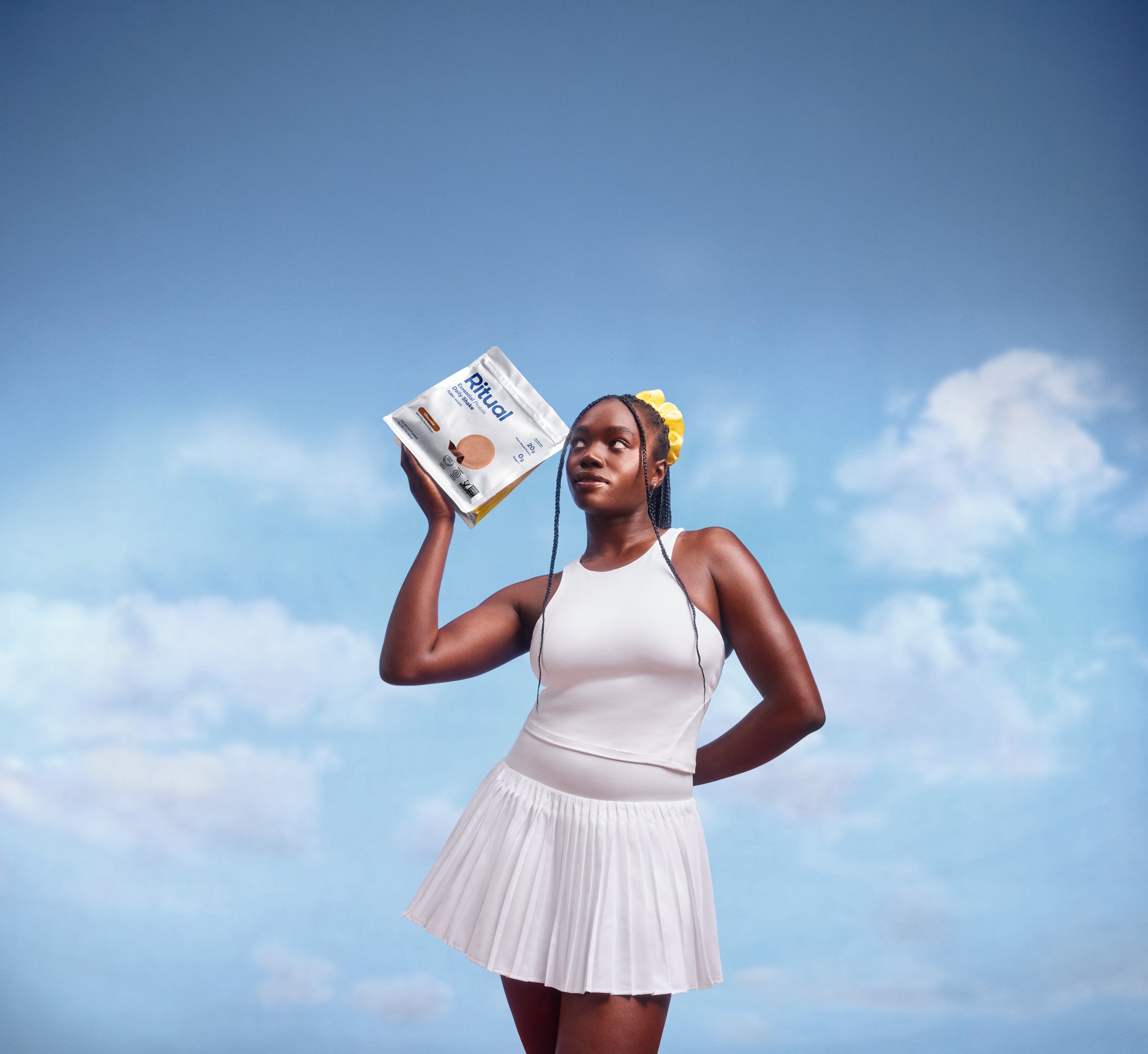 A young woman with braided hair and yellow hair accessories holding a packet of Ritual protein powder against a blue sky background.