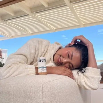 A woman smiling and resting her head on her arm on a white surface, with a bottle of Irem spray next to her, under a white slatted roof with a blue sky in the background. Hair and makeup by Amber Rose.