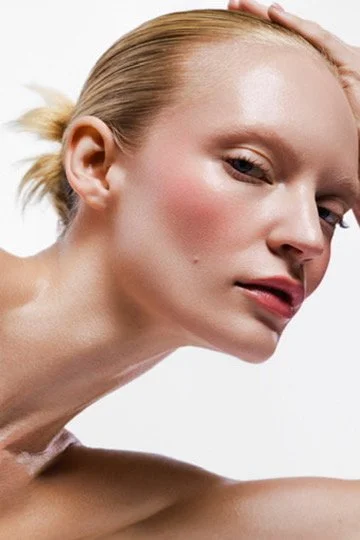 Close-up of a young woman with blonde hair, fair skin, and natural makeup, touching her head with her right hand. Hair and makeup by Amber Rose.