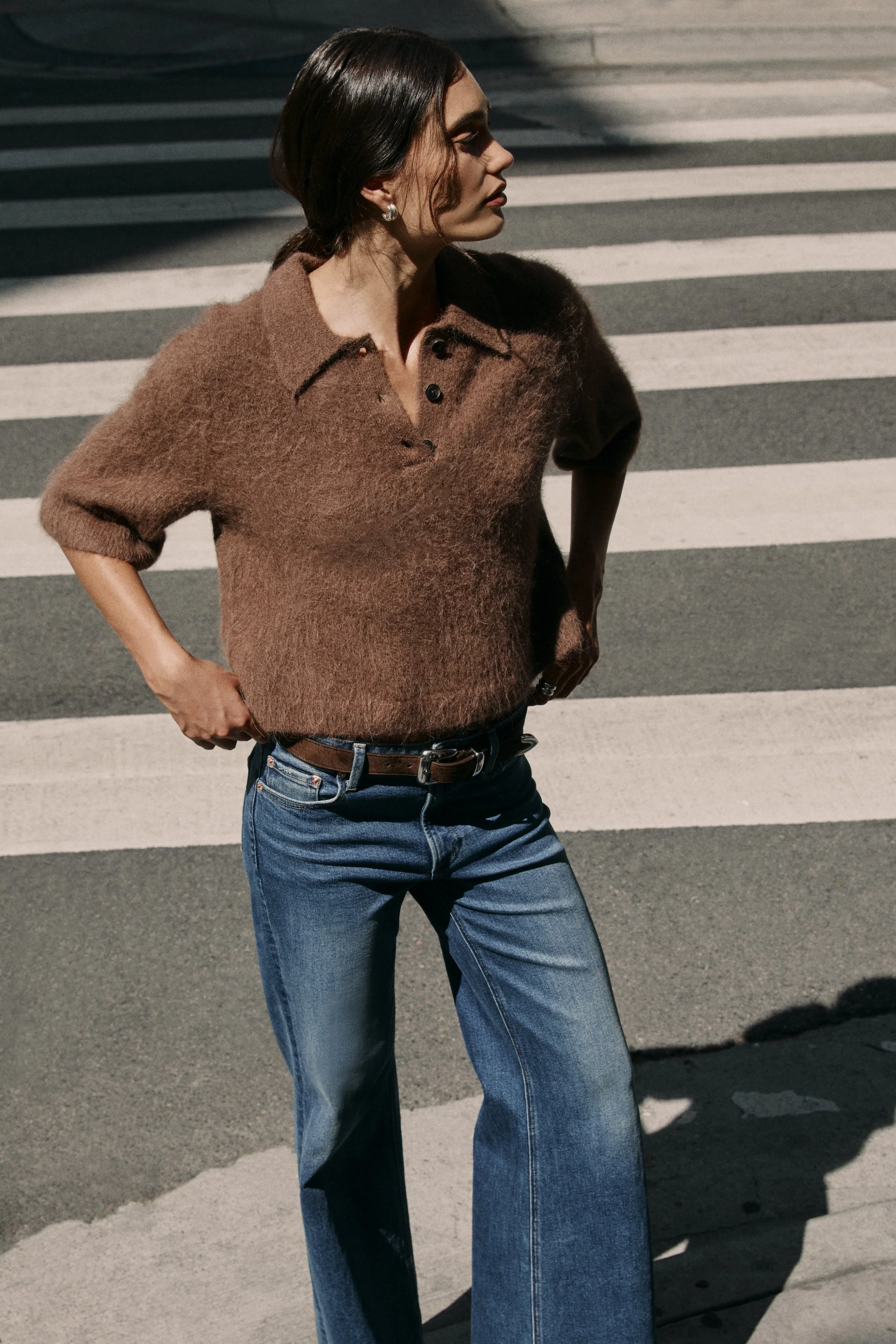 A woman with dark hair and earrings is crossing the street on a pedestrian crosswalk, wearing a brown sweater and blue jeans, with her hands on her hips.