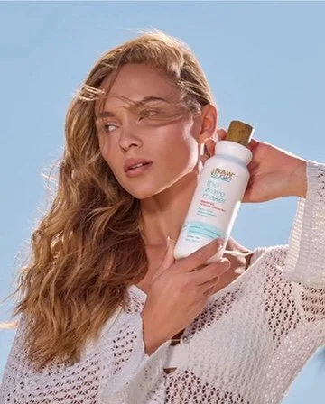A woman with wavy blonde hair holds a bottle of sunscreen near her face, outdoors on a sunny day.